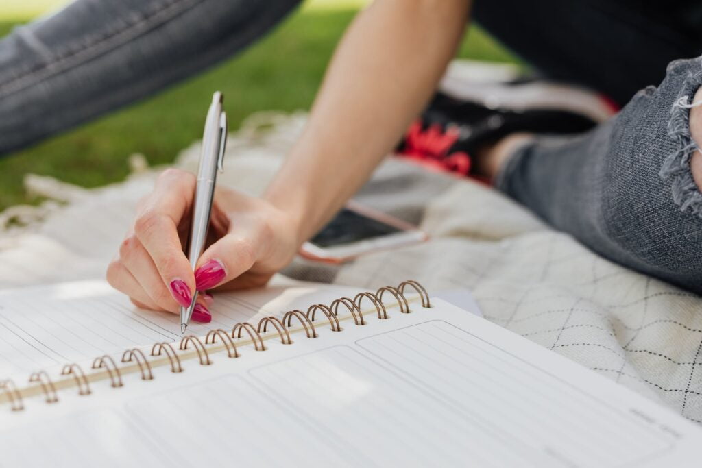 Crop anonymous lady in casual wear writing down plans for next day in organizer while sitting on blanket in quiet summer park