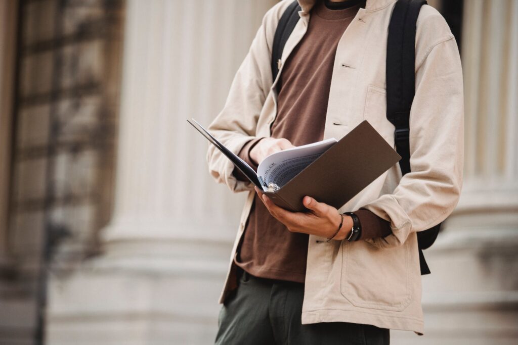 Crop anonymous male learner with paper files standing near building of university with columns